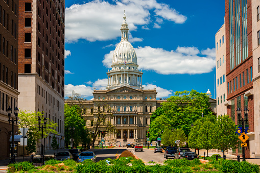 Lansing, United States - May 24, 2014 - The Michigan State Capitol as viewed from within Downtown Lansing, with trees, plants, office buildings, and pedestrians and cars and a driver in the foreground, and a blue sky with clouds in the background.