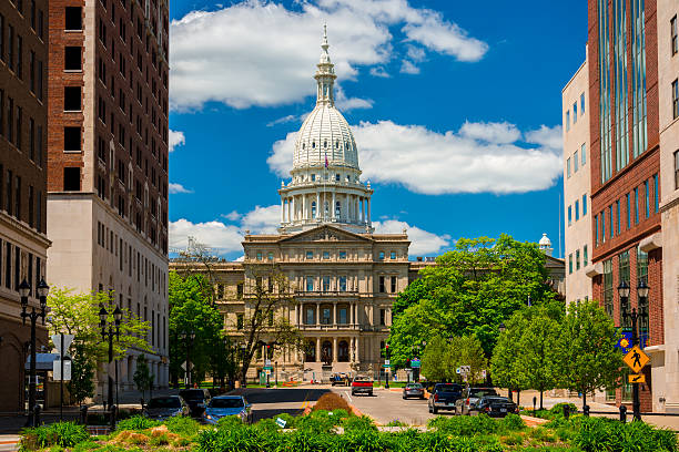 Michigan State Capitol and Downtown Lansing Lansing, United States - May 24, 2014 - The Michigan State Capitol as viewed from within Downtown Lansing, with trees, plants, office buildings, and pedestrians and cars and a driver in the foreground, and a blue sky with clouds in the background. Michigan stock pictures, royalty-free photos & images