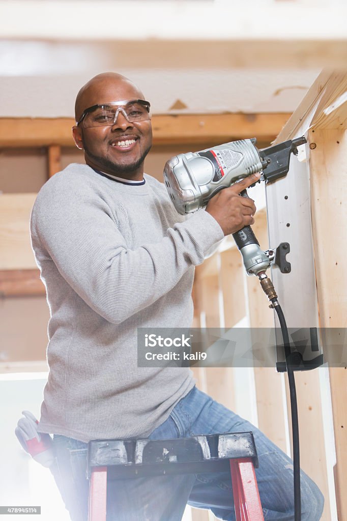 Black construction worker with nail gun A construction worker, an African American man in his 40s, working on a home remodeling project.  He is standing on a ladder with a nail gun, nailing wood posts.  He is smiling at the camera, wearing safety glasses. 40-49 Years Stock Photo