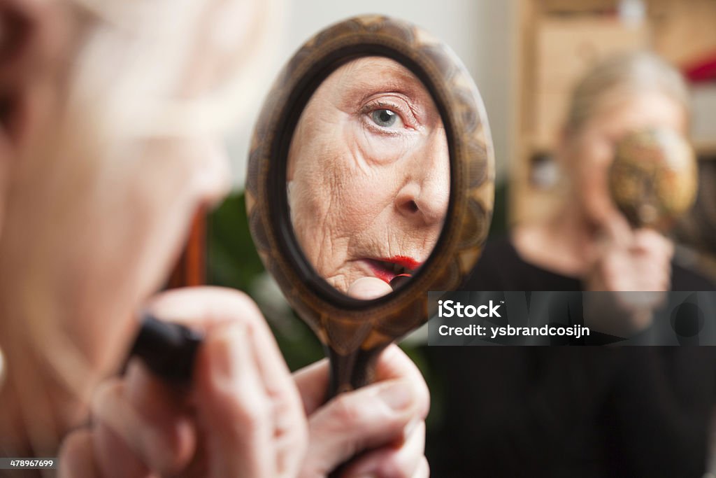 Good looking senior woman doing lipstick in front of mirror. Good looking senior woman doing make-up in front of mirror. Senior Women Stock Photo