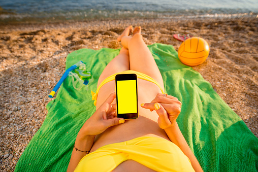 Young woman in swimsuit using mobile phone with empty screen for copy paste lying on the green towel on the beach. Top view focused on the hand with phone.