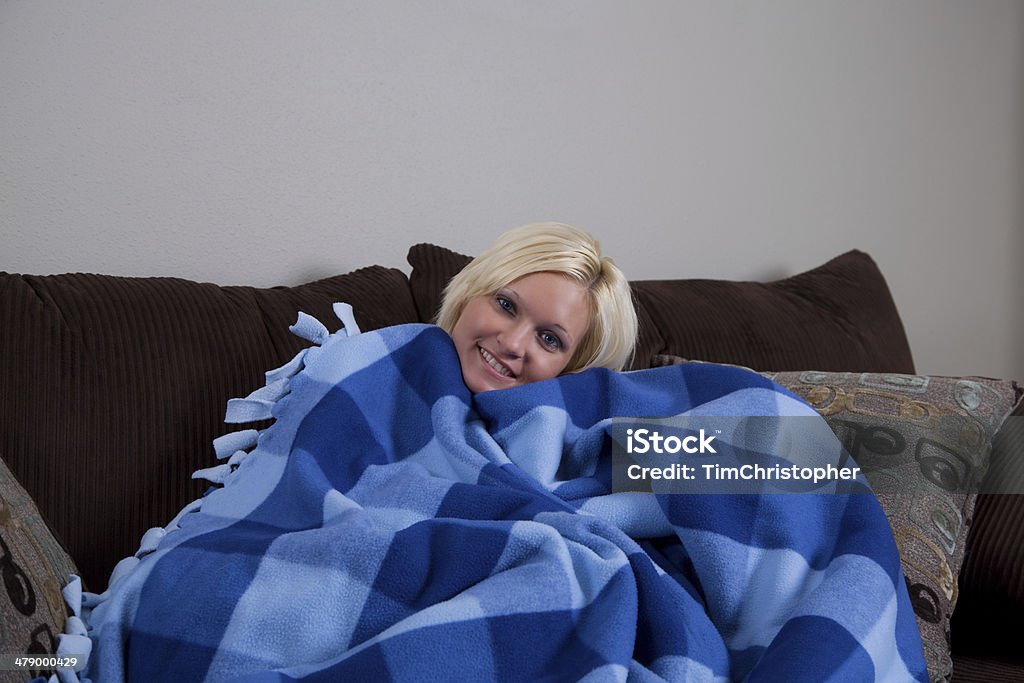 Relaxing and Staying Warm on Couch A blond woman is covered by a warm blanket, sitting on a couch. Adult Stock Photo