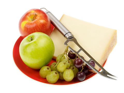a DSLR royalty free image of a plate of Beaufort cheese with a selection of fresh ripe juicy fruit including a small bunch of red and green grapes and a single red and green apple with a cheese knife, isolated against a plain white background
