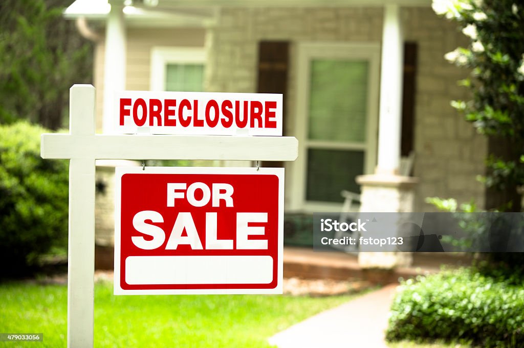 Foreclosure, house for sale sign. Front yard of home. Nobody. Red and white "Foreclosure, Home for Sale" sign in front of a stone, wood house that is for sale and is being foreclosed upon by a financial institution. Green grass and bushes indicate the spring or summer season. Front porch and windows in background.  Economic depression, recession, bankruptcy concepts. Foreclosure Stock Photo