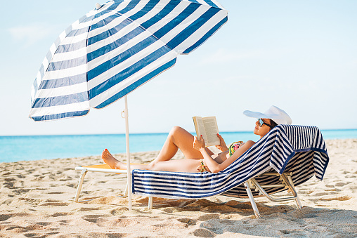 Beautiful young woman in bikini lying at the beach, reading a book and suntanning on sunny day. Woman wearing sunglasses and white hat near the seaside in summer. Image taken with Nikon D800 and 24-70 lens in XXXL size, developed from RAW file