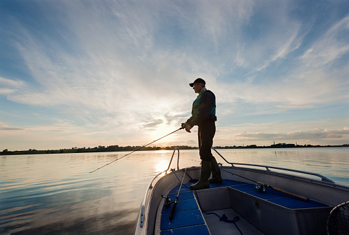 Portrait of a 65 year old  fisherman waiting patiently for a fish to bite whilst standing on his boat  fishing on a flat calm sea. Horizontal format photographed against a setting sun with lots of copy space in the sky. Photographed on Stege Nor on the island of Møn or Moen in Denmark.