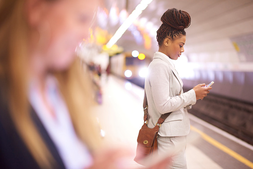 A young businesswoman is standing at an underground subway station checking her phone. In the background a tube train is approaching with it's lights lighting the inside of the tube . In the foreground another woman is shown defocussed .