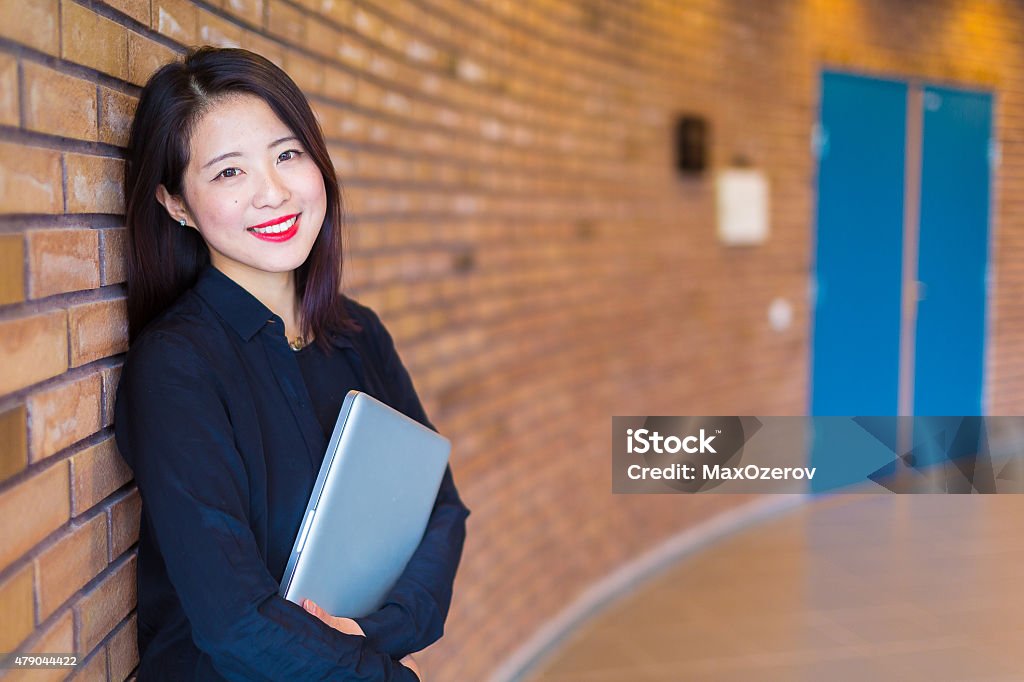 Asian college student standing at the brick wall with laptop A portrait of an Asian college or university student at campus. She is standing at a red brick wall holding her laptop. University Student Stock Photo