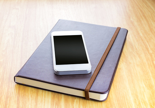 Blank screen Smartphone on Brown Hard cover notebook with elastic strap on wooden table in perspective view,Template for adding your content.