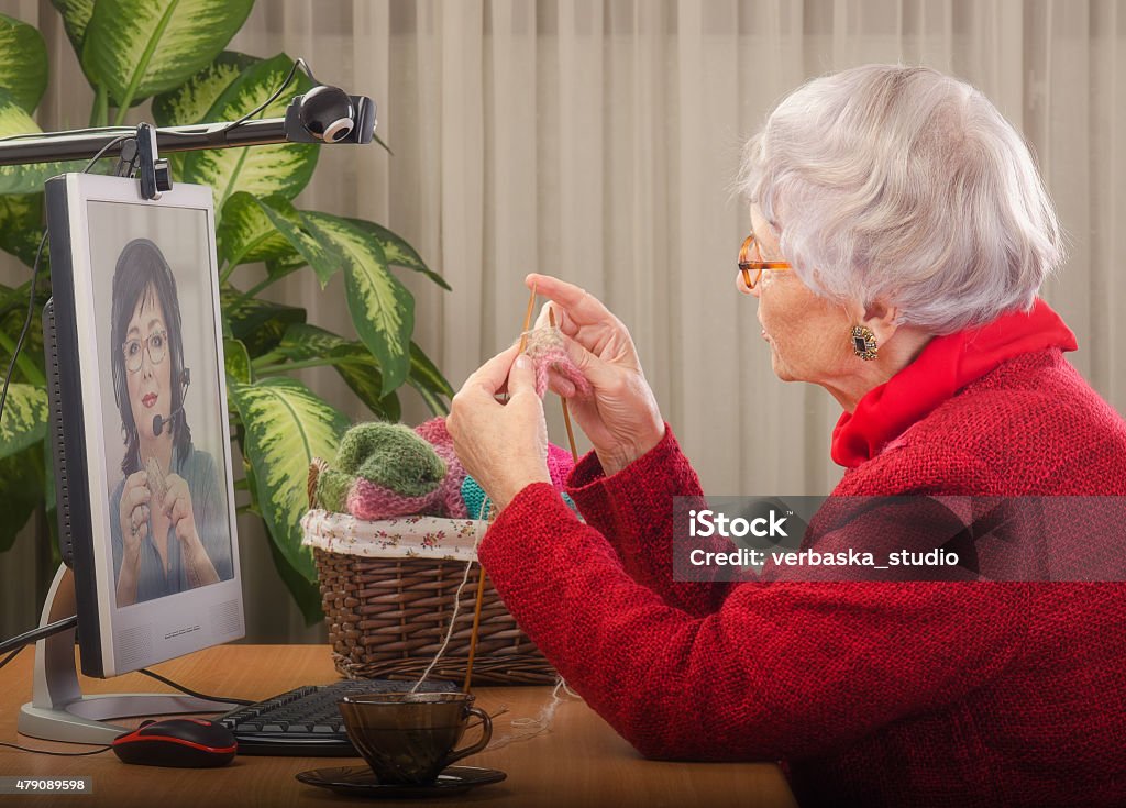 First steps of learning to knit online Well-trained private teacher is explaining to online learner how to knit.  Senior woman is knitting in front of desktop. Two webcams are carefully tracing her work for staring student in monitor. Side shot 40-49 Years Stock Photo