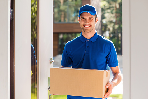 Cheerful young courier holding a cardboard box while standing at the entrance of apartment