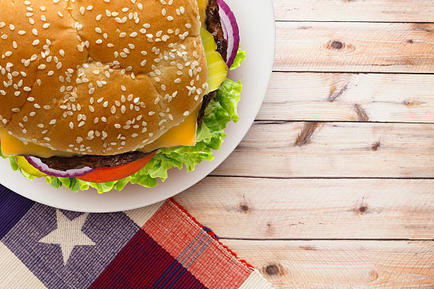 Plated cheese burger on patriotic theme napkin with lettuce, tomatoes, pickles and onions on a rustic picnic table.