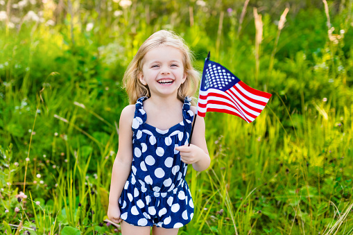 Beautiful little girl with long curly blond hair with american flag in her hand laughing on sunny day in summer park. Independence Day, Flag Day concept
