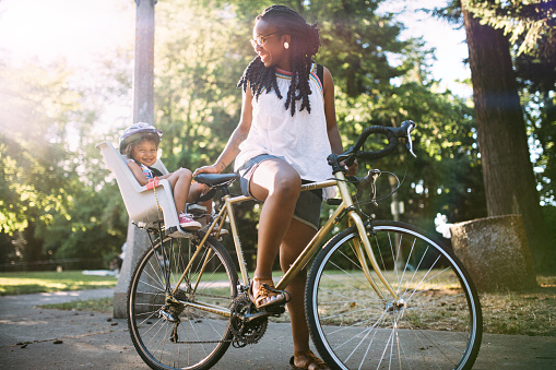 A mother and her kindergarten aged girl smile lovingly as they take a break on a bike trip in the park. The daughter sits safely in a children's bike seat.  Image meant to emphasize mother and daughter relationship as well as outdoor activities and exercise.  Horizontal image.
