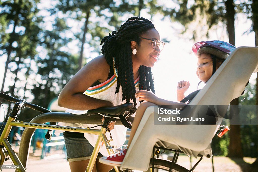 Mom Buckles Up Daughter in Bike A mother and her kindergarten aged girl smile lovingly as mom buckles her daughter into a children's bike seat for a bicycle ride together through the park.  The girl wears a fun stylish helmet.  Image meant to emphasize mother and daughter relationship as well as outdoor activities and exercise.  Horizontal image. Child Stock Photo