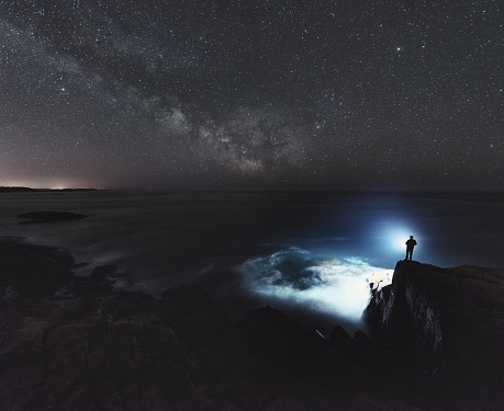 A man is silhouetted against the light of a bright spotlight as he searches the Atlantic under the Summer Milky Way.  Long exposure.