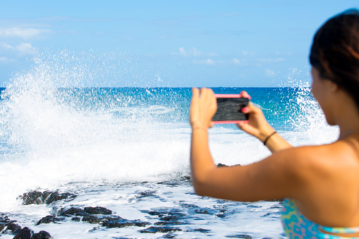 Young woman taking a picture of breaking waves at Erma's on Oahu, Hawaii in summer sunshine