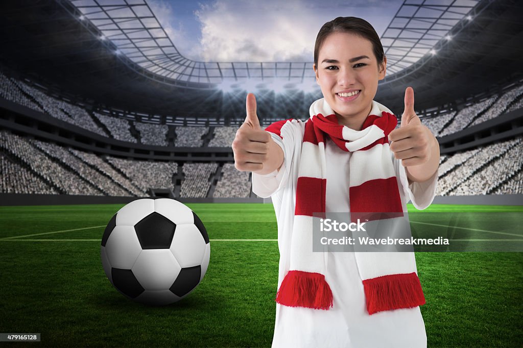 Football fan in white wearing scarf showing thumbs up Football fan in white wearing scarf showing thumbs up against vast football stadium with fans in white Scarf Stock Photo