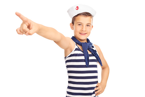 Studio shot of a little male kid in a sailor costume pointing forward with his hand and looking at the camera isolated on white background