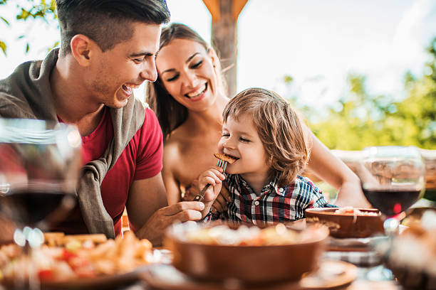 Cheerful young father feeding his son in restaurant. Happy young parents enjoying with their little boy while having a lunch on restaurant terrace. Father is feeding son who is sitting on mothers lap. kids restaurant pictures stock pictures, royalty-free photos & images
