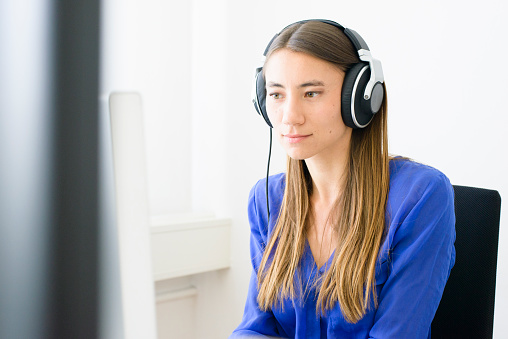 A young woman in a modern office, wearing headphones, working with a computer. She has long hair and is wearing a blue blouse. Camera: 36MP Nikon D800E.