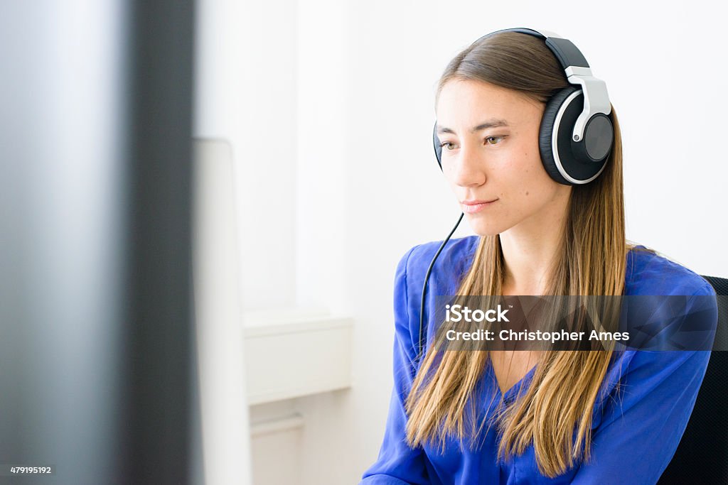 Young Professional Woman Wearing Headphones Concentrating on Computer Screen A young woman in a modern office, wearing headphones, working with a computer. She has long hair and is wearing a blue blouse. Camera: 36MP Nikon D800E. 2015 Stock Photo