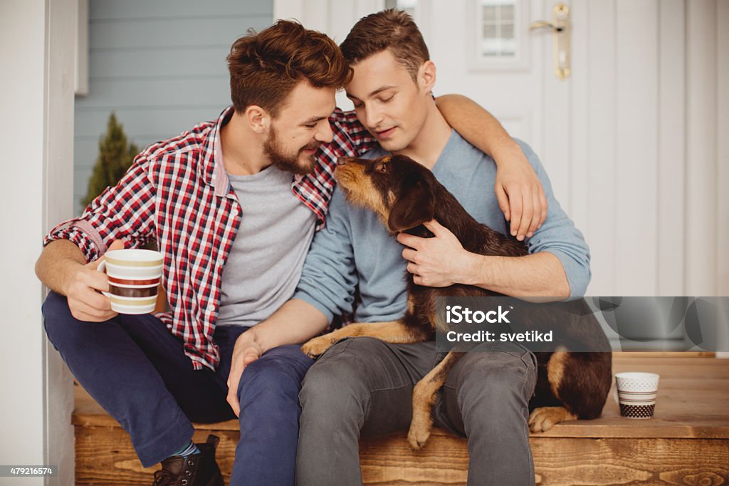 Young gay couple. Young gay couple sitting on porch in front of their home with their dog. Drinking coffee. Caucasian ethnicity, blond hair, casual. Dog Stock Photo