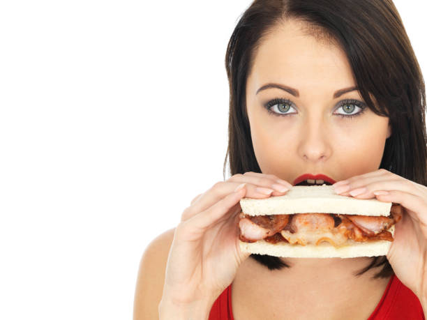 Attractive young woman, with dark straight mid length hair, eating a crispy grilled bacon sandwich on white sliced bread, holding the bacon sandwich straight on in front of the camera, wearing a red vest top, looking into the camera, isolated on a white plain background, clipping path, cut out, room for text or copy