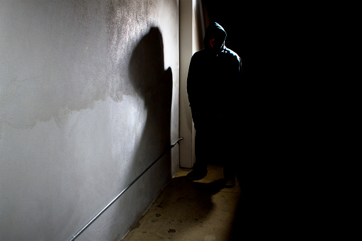 Photo of a hooded criminal stalking in the shadows of a dark street alley.  The hooded man is a silhouette and hiding in the dark.  The man is a criminal waiting to ambush victims.  The concrete walls provide copyspace.The photo depicts crime.