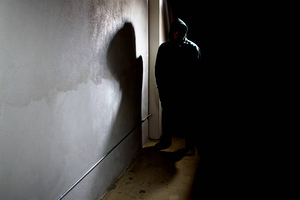 Stalker in a Dark Alley Photo of a hooded criminal stalking in the shadows of a dark street alley.  The hooded man is a silhouette and hiding in the dark.  The man is a criminal waiting to ambush victims.  The concrete walls provide copyspace.The photo depicts crime. hiding place stock pictures, royalty-free photos & images