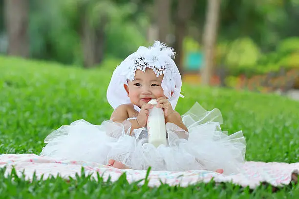 Little asian girl wearing a white dress and sucking the milk bottle sitting on the grass.