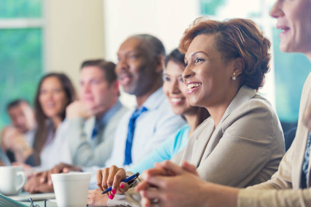 African American businesswoman listening to seminar speaker at business conference Mid adult African American businesswoman is smiling while attending business conference. She is listening to speaker in seminar while she sits in a row with other diverse professional businesspeople. Woman is wearing business casual clothing and is taking notes. She's drinking a cup of coffee. board room stock pictures, royalty-free photos & images