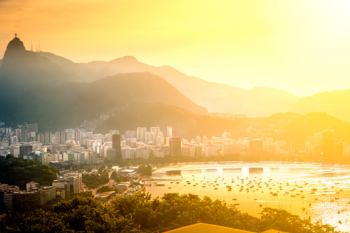 Aerial view of Rio de Janeiro at sunset.  The beach is visible in the bottom-right section of the photo, and several boats dot the water.  There is a small forested area in the middle of the photo.  Buildings surround the beach and forest area, extending toward the mountains in the background.  The mountains are blanketed by mist that partially obscures them, but the Christ the Redeemer statue is visible above the mist.  The sunlight adds a warm glow to the photo, and the area behind the mountains is golden.  