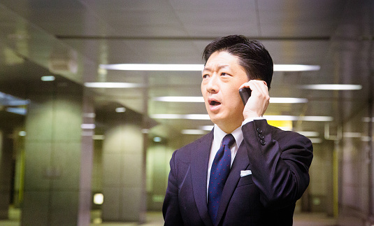 Stressed Japanese businessman shocked by terrible news on his mobile phone. He is wearing a suit and is frowning, mouth open, in complete disbelief. Shot underground as he commutes to work.