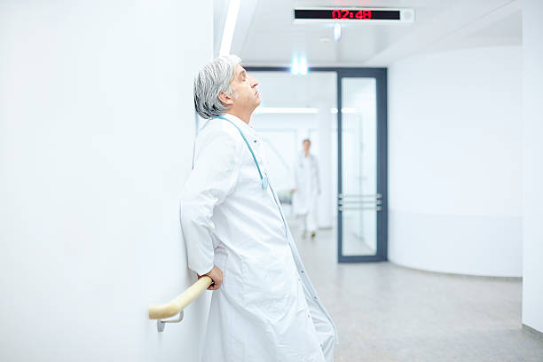 Tired and stressed female doctor in white coat and blue scrubs standing outside hospital, touching her forehead and feeling emotional exhaustion after a long workday.