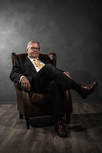 Successful senior businessman in a suit relaxing in an armchair in a shadowy room with grey wall and wooden floor