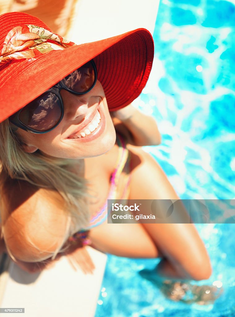 Woman relaxing by swimming pool. Top view of attractive blond woman sitting by poolside. She's smiling with beautiful se of teeth and looking at camera, wearing sunglasses and red sun hat. Her legs are crossed. 20-29 Years Stock Photo