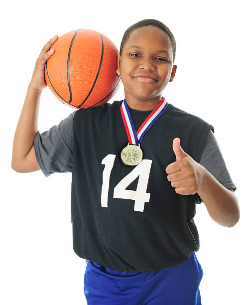 A happy preteen athlete supporting his basketball on his shoulder, a thumbs up with the other hand and a red, white and blue winning ribbon and metal around his neck.  On a white background.