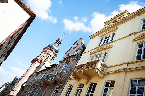 View along rowhouses and buildings in historical center of Heidelberg in summer. In background is a belfry of church.