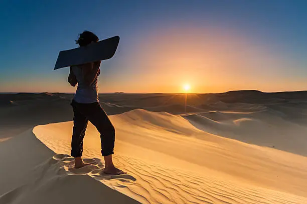 Young female sandboarding in The Sahara Desert. She is standing on a sand dune, holding sandboard and watching the sunse over desert. The Sahara Desert is the world's largest hot desert with the biggest sand dunes.http://bem.2be.pl/IS/egypt_380.jpg