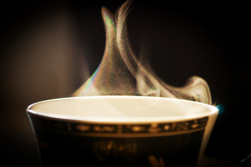 Steam from a coffee/tea cup backlit with bright sunlight and showing a rainbow on the left side of the steam and a flare on the rim of the coffee cup on the right. Focus is on the steam with the cup out of focus.