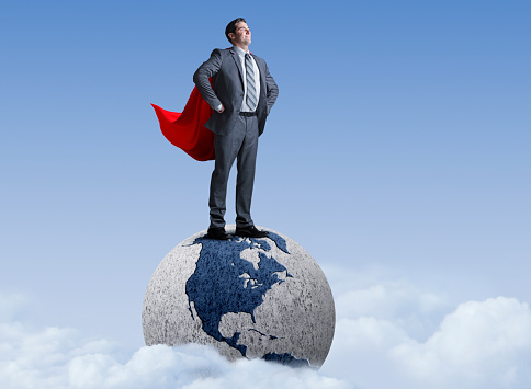 A businessman wearing a red cape stands on top of a globe showing North America.  The globe emerges from clods and stands out against a blue sky.