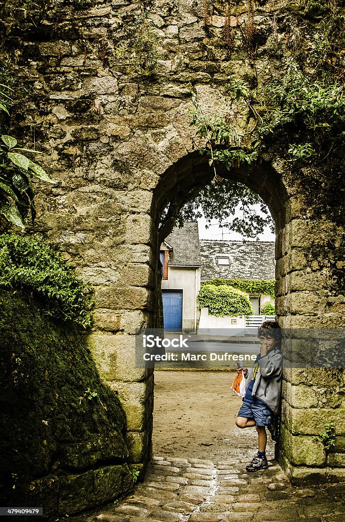 Kid in a arch door Young boy, eating, standing and leaning on the side of a big stone arch door with plants growing all on the stone Brittany - France Stock Photo