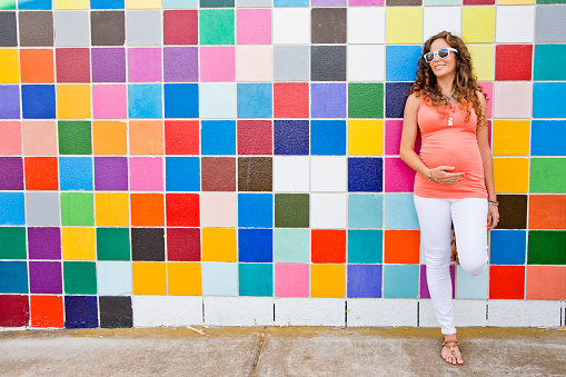 Candid maternity photo in front of a colourful wall. 7 months pregnant. Curly-haired pregnant woman smiling and wearing matching sunglasses.