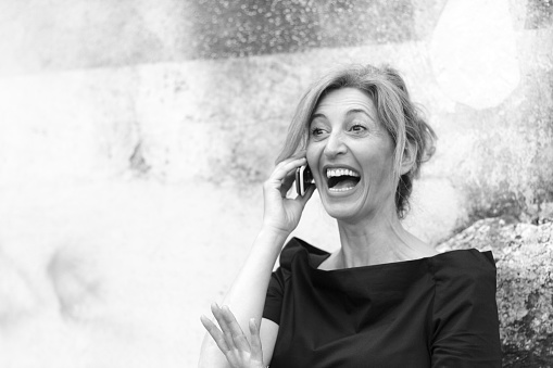 Blonde, 40s, speaking on the phone looking very amused/delighted/urprised.  B&W, with copy space on the old stone wall behind her. Shot in Sicily, Italy, with a Sicilian model.