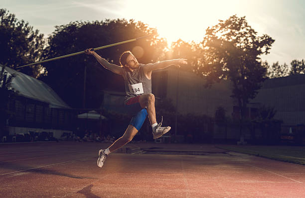 Athlete training for javelin throw on sports track with blue sky background. Sports equipment and green field visible around illustrating sports environment