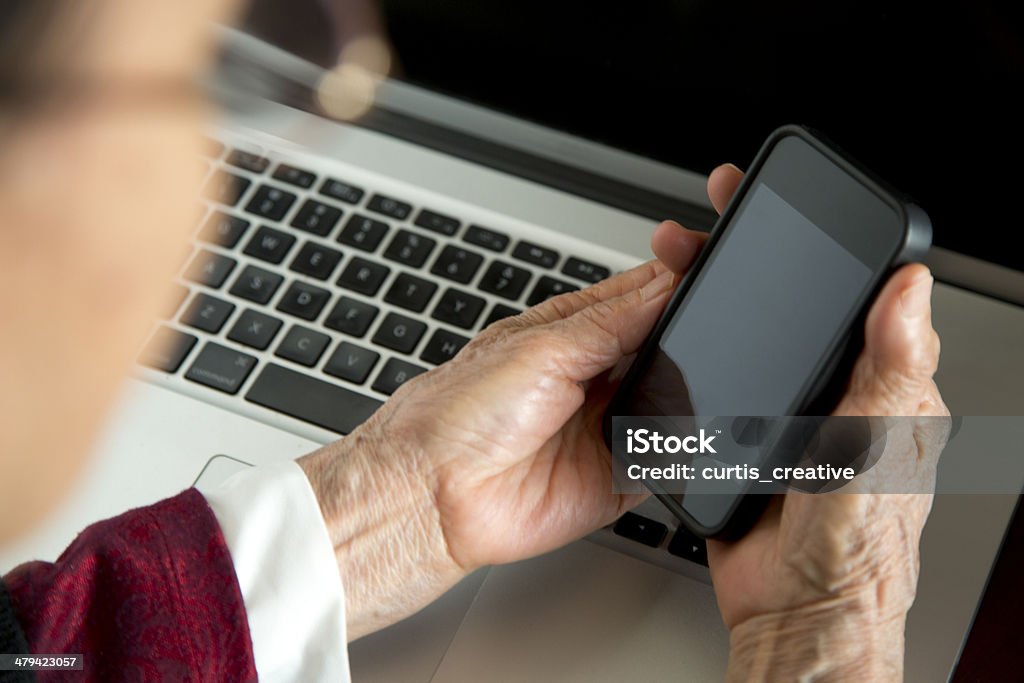 High tech grandma with her mobile phone Woman in her 80's holding mobile phone over her laptop computer. 80-89 Years Stock Photo