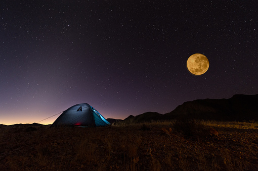 A full, bloody moon rising over a desert landscape and tent lit from inside. Stars visible. Shot in the Northern Cape of South Africa. 