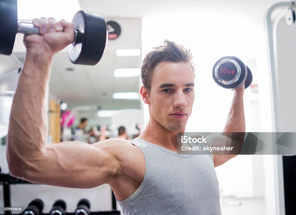 Young man exercising with dumbbells and looking at camera. Serious young man during weight lifting training in a gym looking at camera. 2015 Stock Photo