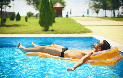 Closeup of handsome caucasian guy relaxing on yellow colored airbed while floating in swimming pool. Rear view. The guy has his arms outstretched and touching the water.